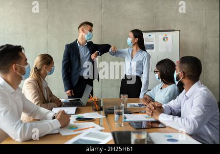 Comunicazione aziendale durante Pandemic. Colleghi in maschere mediche urtare i gomiti in ufficio Foto Stock