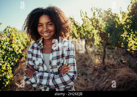 Razza mista femmina contadina in piedi con braccia incrociate sorridenti in piedi in splendidi vigneti Foto Stock