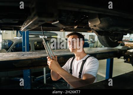 Mechanic professionale dell'automobile maschio messo a fuoco in piedi uniforme nella fossa di ispezione e lavorando con l'attrezzo. Foto Stock