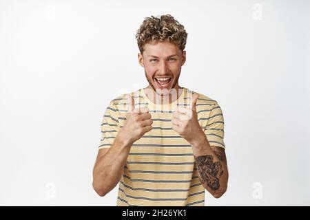 Felice ragazzo biondo ridendo e sorridendo, mostrando i pollici in su per approvazione, come qualcosa di buono, dicendo sì, in piedi su sfondo bianco Foto Stock