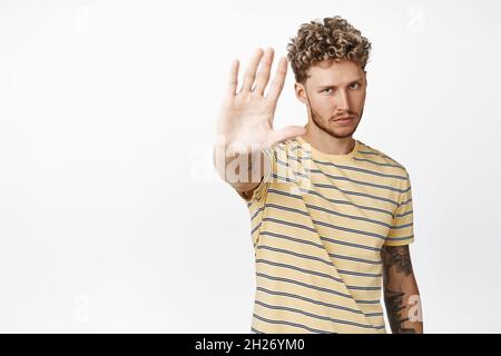 L'uomo curly biondo serio allunga la mano e guarda sicuro, dicendo di smettere, dicendo no, proibisci l'azione, in piedi in maglietta su sfondo bianco Foto Stock