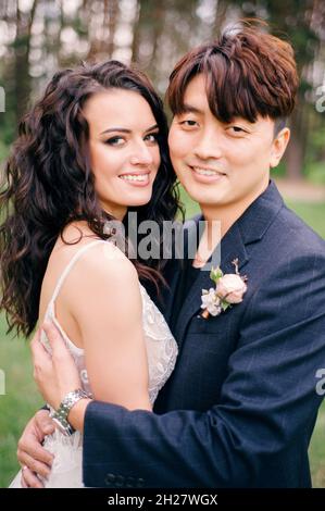 Coppia internazionale. Giorno del matrimonio. Giovani sposi novelli felici nella foresta verde fresca. Ritratto di sposa e sposo. Matrimonio di primavera. Cerimonia nuziale all'aperto. Foto Stock