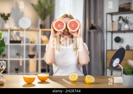 Donna caucasica in t-shirt bianca che copre gli occhi sotto fette di pompelmo fresco. Felice bionda che passa il tempo a casa per le procedure di bellezza. Concetto di cura della pelle. Foto Stock