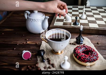 la mano tiene un pezzo bianco vintage di scacchi. Scacchiera e scacchi sparsi su tavola di legno. Ciambella al cioccolato per colazione al mattino. Concetto di gioco da tavolo Foto Stock