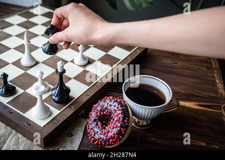 la mano tiene un pezzo bianco vintage di scacchi. Scacchiera e scacchi sparsi su tavola di legno. Ciambella al cioccolato per colazione al mattino. Concetto di gioco da tavolo Foto Stock