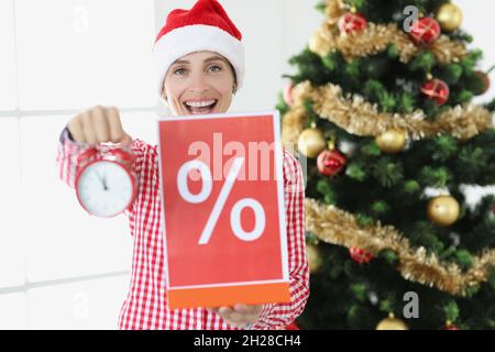 Giovane donna sorridente in babbo natale cappello tiene sveglia e poster con la percentuale sullo sfondo di albero di Capodanno Foto Stock
