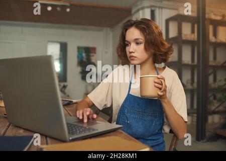 Donna seria proprietario di piccola impresa seduto al tavolo e utilizzando il computer portatile, mentre beve caffè e di lavoro in ceramica officina Foto Stock