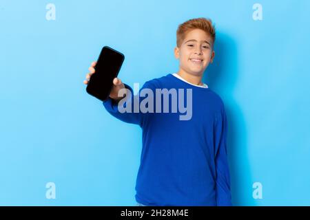 Ragazzo con capelli rossi in pullover casual che mostra smartphone isolato su sfondo blu. Foto Stock