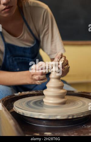 Primo piano di una ragazza irriconoscibile in grembiule che scolpisce il bel vaso di argilla con le mani sporche sulla ruota in ceramica Foto Stock
