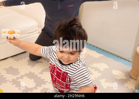 bambino di 2 anni a casa volare giocattoli aeroplano nelle sue mani, padre in background Foto Stock