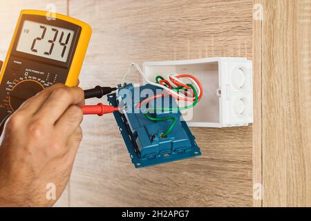 Un elettricista che utilizza un misuratore digitale per misurare la tensione su una presa a muro su una parete di legno. Foto Stock