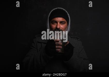 Uomo senza tetto di mezza età con una barba grigia che tiene una tazza di tè caldo per riscaldarsi in una notte fredda Foto Stock