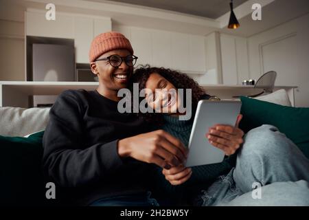 Coppia di adulti alla moda, multi-etnico, ridendo a video su tablet sul divano in appartamento moderno Foto Stock