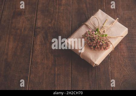 Confezionato in casa presente in carta kraft e fiori rosa su un tavolo di legno. Immagine ravvicinata di una bella scatola regalo decorata con fiori Foto Stock