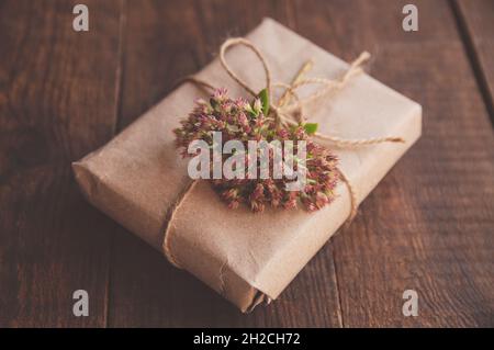 Confezionato in casa presente in carta kraft e fiori rosa su un tavolo di legno. Immagine ravvicinata di una bella scatola regalo decorata con fiori Foto Stock