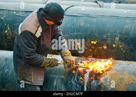 La saldatrice taglia grandi tubi metallici con saldatura in ocetilene. Un lavoratore in strada taglia tubi di grande diametro durante il giorno e scintille e fuoco volare. Foto Stock