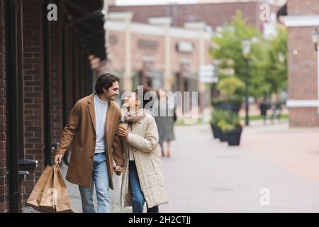 gioiosa coppia multietnica in cappotti che tengono le mani mentre si guarda l'un l'altro e camminare con gli acquisti Foto Stock