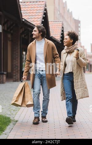 gioiosa coppia multietnica in cappotti che tengono le mani mentre si cammina con acquisti vicino al centro commerciale Foto Stock