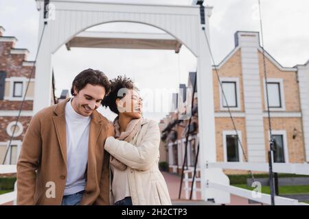felice coppia multietnica in cappotti ridendo vicino al centro commerciale Foto Stock