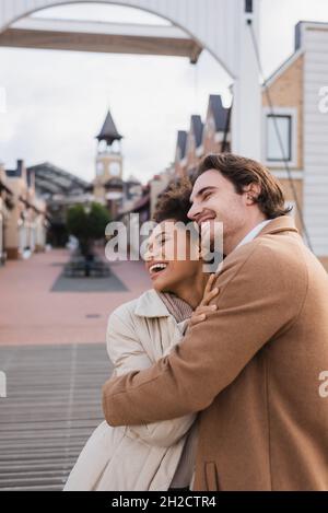 gioiosa coppia multietnica in cappotti che abbracciano vicino al centro commerciale Foto Stock