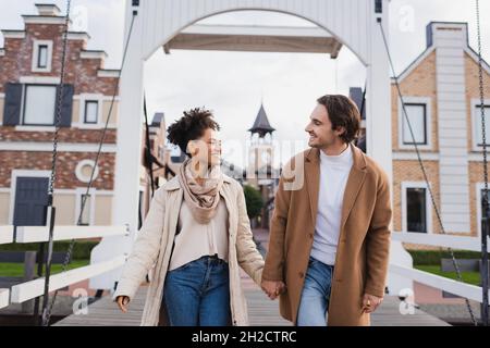 sorridente coppia multietnica in cappotti che tengono le mani vicino al centro commerciale Foto Stock