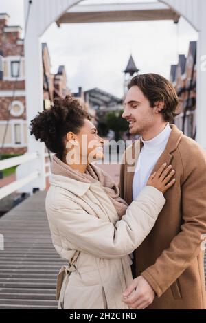 sorridente coppia multietnica in cappotti che abbracciano vicino al centro commerciale Foto Stock