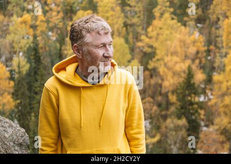 Un autentico ritratto di un sorridente viaggiatore di mezza età non rasato in una felpa con cappuccio gialla sulla cima di una montagna sullo sfondo di una foresta autunnale. Foto Stock