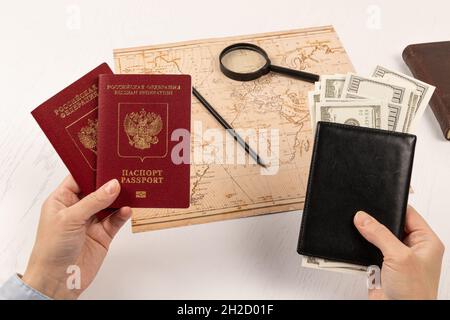 le mani femminili tengono due passaporti e un portafoglio in pelle nera con soldi. su un tavolo di legno bianco si trova una mappa del mondo, una lente d'ingrandimento, una matita, Foto Stock