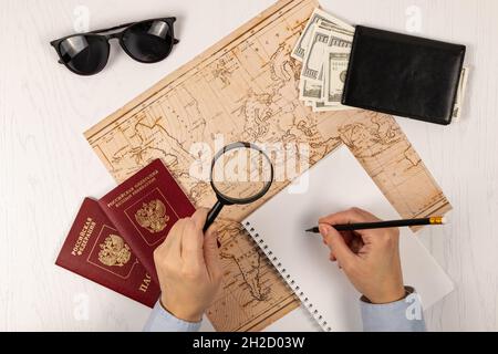 la mano femminile fa note con una matita in un taccuino. su un tavolo di legno bianco si trova una mappa del mondo, due passaporti, una lente d'ingrandimento e una wa nera Foto Stock