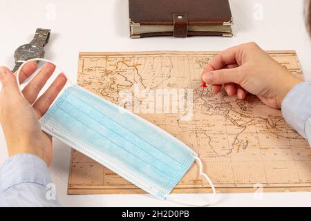 la mano femminile mostra il luogo di viaggio sulla mappa del mondo. su un tavolo di legno bianco una maschera medica blu, un diario e un orologio da polso. primo piano Foto Stock