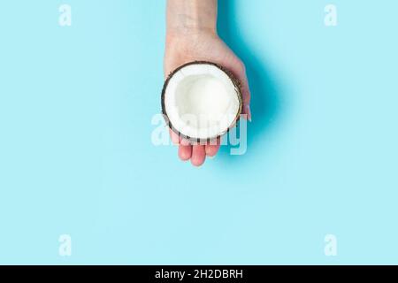 La palma di una donna tiene una metà aperta di una noce di cocco su uno sfondo blu. Vista dall'alto, disposizione piatta. Foto Stock