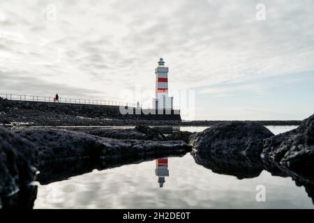 Splendida vista della torre faro di colori rosso e bianco sulla riva rocciosa sotto il cielo nuvoloso in Islanda Foto Stock