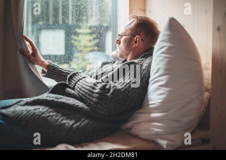 Ritratto di pensiero triste uomo di mezza età in occhiali vestito cardigan aperto giacente su letto accogliente accanto alla finestra che guarda fuori strada attraverso vetro raindrop Foto Stock