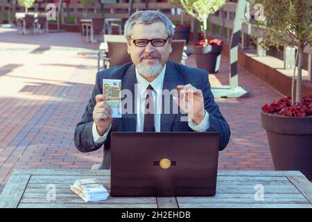 L'uomo finanziatore tiene soldi nelle sue mani e bitcoins. Ha fatto soldi con il suo laptop che commercia sulla borsa. Foto Stock