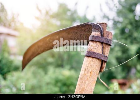 Vecchio scythe arrugginito su manico di legno su sfondo sfocato con effetto bokeh nel cortile di casa di campagna in estate closeup estremo Foto Stock