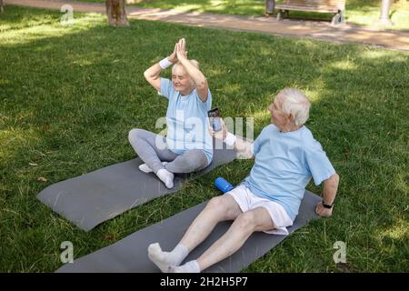 L'uomo anziano spara la moglie che fa yoga asana sull'erba del campo nel parco Foto Stock