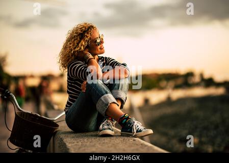 Donna hipster con capelli ricci e occhiali da sole seduti sul muro di argine e ammirando la bella natura con la bicicletta parcheggiata contro il muro. Femmina tou Foto Stock