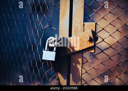 Un vecchio cancello arrugginito con una griglia di metallo strappato è chiuso da un lucchetto forte in una giornata di sole. Territorio chiuso. Foto Stock