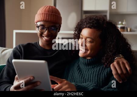 Coppia di adulti alla moda, multietnico, sorridente al tablet sul divano in appartamento moderno Foto Stock