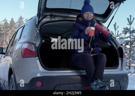 Donna anziana che beve il tè mentre si siede sul retro del suv dopo la strada della foresta di viteria. Attività all'aperto per aumentare l'umore durante la pandemia di coronavirus alla pinta Foto Stock