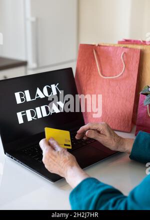 Le mani di una donna anziana stanno tenendo una carta di credito davanti a un monitor portatile con l'iscrizione Black Friday. Foto Stock