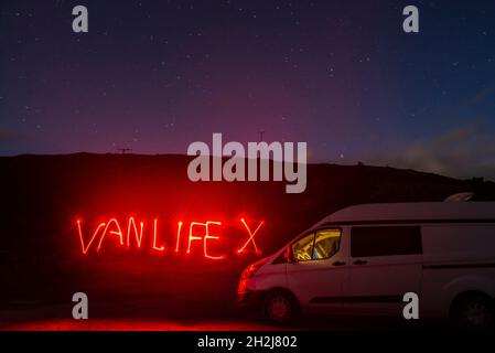 Una lunga esposizione di un campervan parcheggiato di notte con la parola Vanlife scritta in luce rossa. Cielo stellato notturno Foto Stock