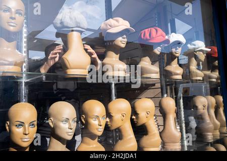 Un negoziante si adatta e regola i cappelli su teste di manichino femminile nella finestra del suo business cappello sulla strada commerciale nel East End, il 21 ottobre 2021, a Londra, Inghilterra. Foto Stock