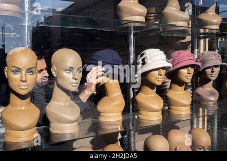 Un negoziante si adatta e regola i cappelli su teste di manichino femminile nella finestra del suo business cappello sulla strada commerciale nel East End, il 21 ottobre 2021, a Londra, Inghilterra. Foto Stock