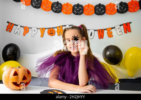 Halloween concetto di vacanza. Ragazza piccola carina in costume da strega seduta dietro un tavolo in Halloween tema camera decorata. Halloween con misure di sicurezza Foto Stock