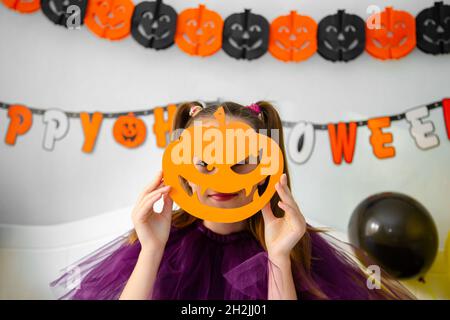 Halloween concetto di vacanza. Ragazza piccola carina in costume da strega seduta dietro un tavolo in Halloween tema camera decorata. Halloween con misure di sicurezza Foto Stock