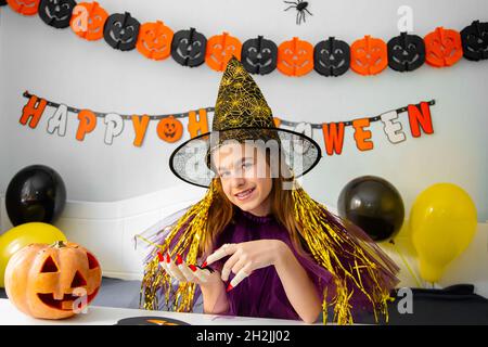 Halloween concetto di vacanza. Ragazza piccola carina in costume da strega seduta dietro un tavolo in Halloween tema camera decorata. Halloween con misure di sicurezza Foto Stock