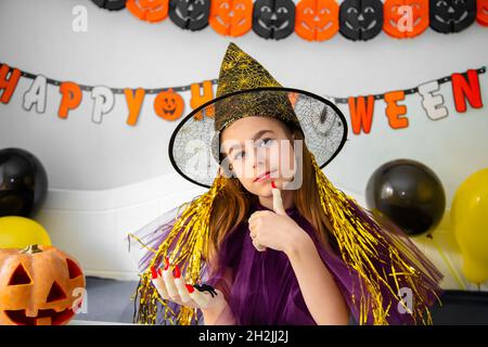 Halloween concetto di vacanza. Ragazza piccola carina in costume da strega seduta dietro un tavolo in Halloween tema camera decorata. Halloween con misure di sicurezza Foto Stock