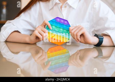 Primo piano corto di una giovane donna irriconoscibile che gioca con il giocattolo arcobaleno pop-it fidget seduto al tavolo. Foto Stock