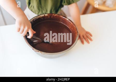 Il bambino che fa la pasta tradizionale al cioccolato per i biscotti di Natale su un tavolo bianco in cucina. Recipiente in metallo e cucchiaio in silicone trasparente. Foto Stock